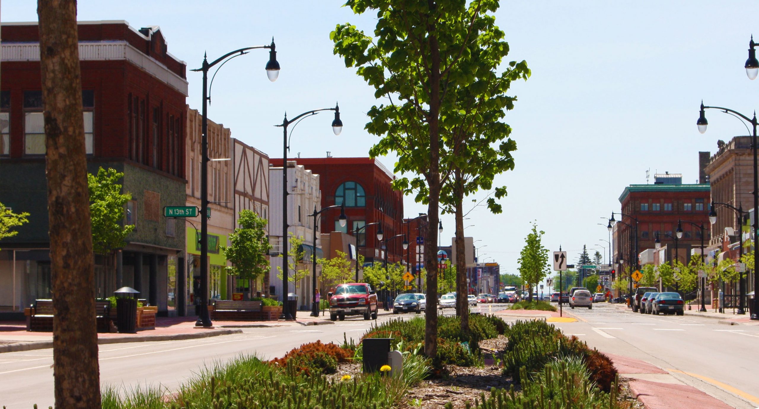 A vibrant row of buildings in the daytime of downtown Superior. There are trees in the middle and lightpoles lining the street.