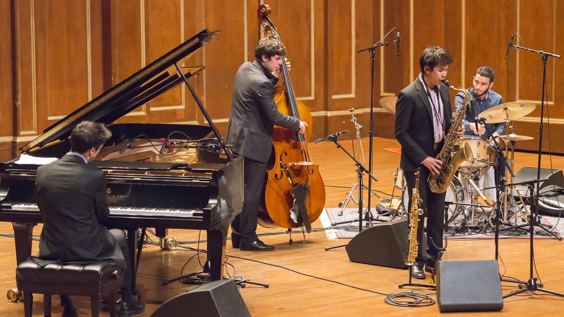 Four male musicians are performing in a jazz ensemble inside a wooden concert hall. One is playing the piano, one is playing the double bass, one is playing the saxophone, and the other is playing the drums.