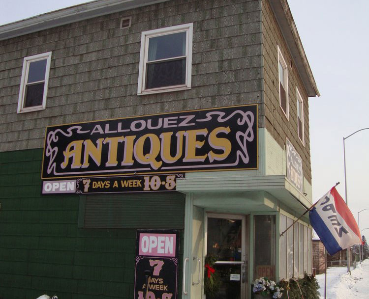 The exterior of an older-style, two story building with an Open flag on the outside. The business sign says Allouez Antiques.