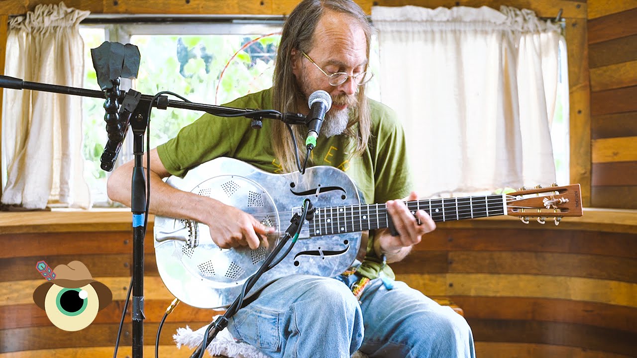 Charlie Parr playing a metal guitar and singing into a microphone. There is a wooden background behind him.