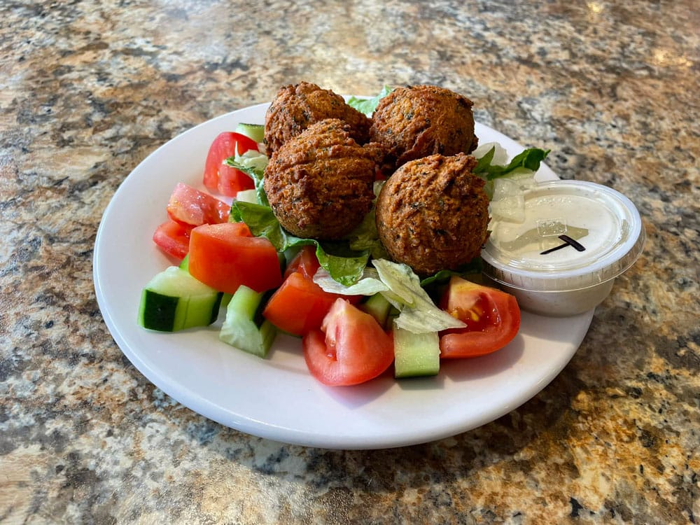A plate of falafel balls with tomatoes, cucumber, lettuce and a white dipping sauce in a little plastic cup.