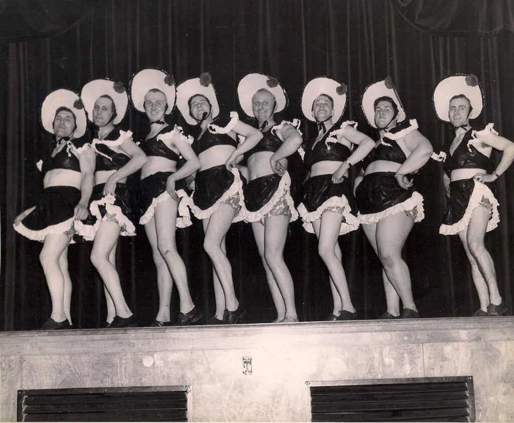 Eight men (yes, men) dressed in black and white ruffled skirts and crop tops, on stage with large bonnets smiling and dancing in a line.