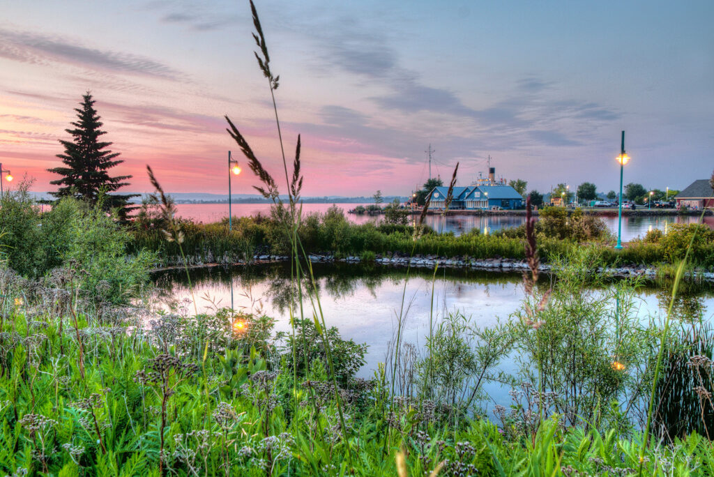 A warm sunset photo of Barker's Island with a pond in the foreground and blue buildings in the distance.