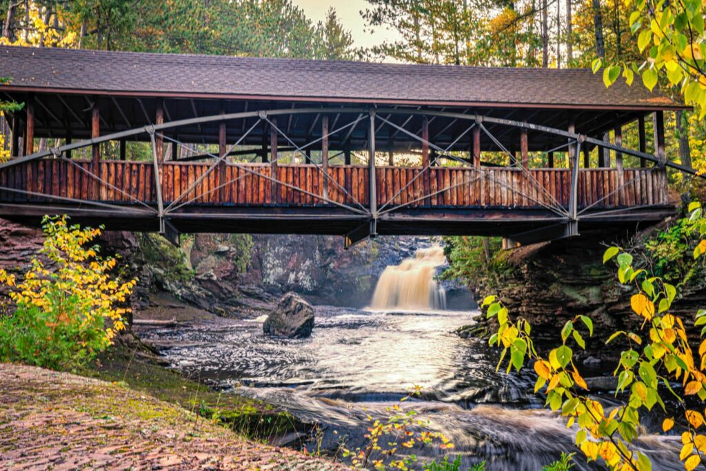 A covered bridge made of logs and arched steel over a river with a cascading waterfall and large rock.