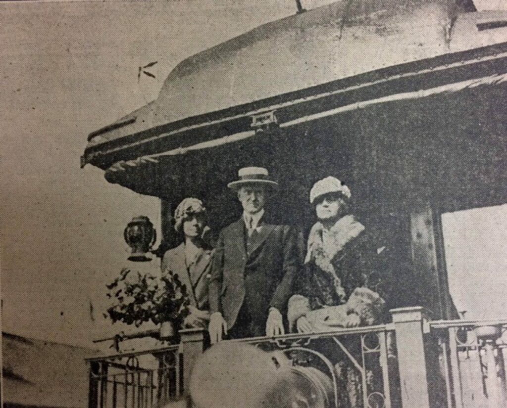 An old, black and white photo of Mr and Mrs Calvin Coolidge and one other woman on the back porch of an old train. The are dressed in heavy business attire and all are wearing appropriate hats for the 1920s.