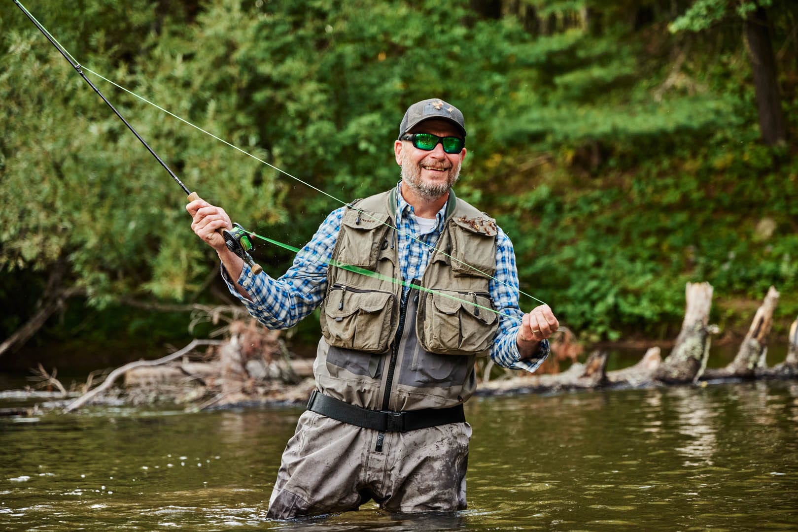 A smiling man with green-tinted sunglasses is fly-fishing in a deep, dark river. He is wearing waders that allow him to be in the water without getting wet. The water is at his groin level. The trees along the river are bright green and there are quite a few tree trunks fallen in the water.