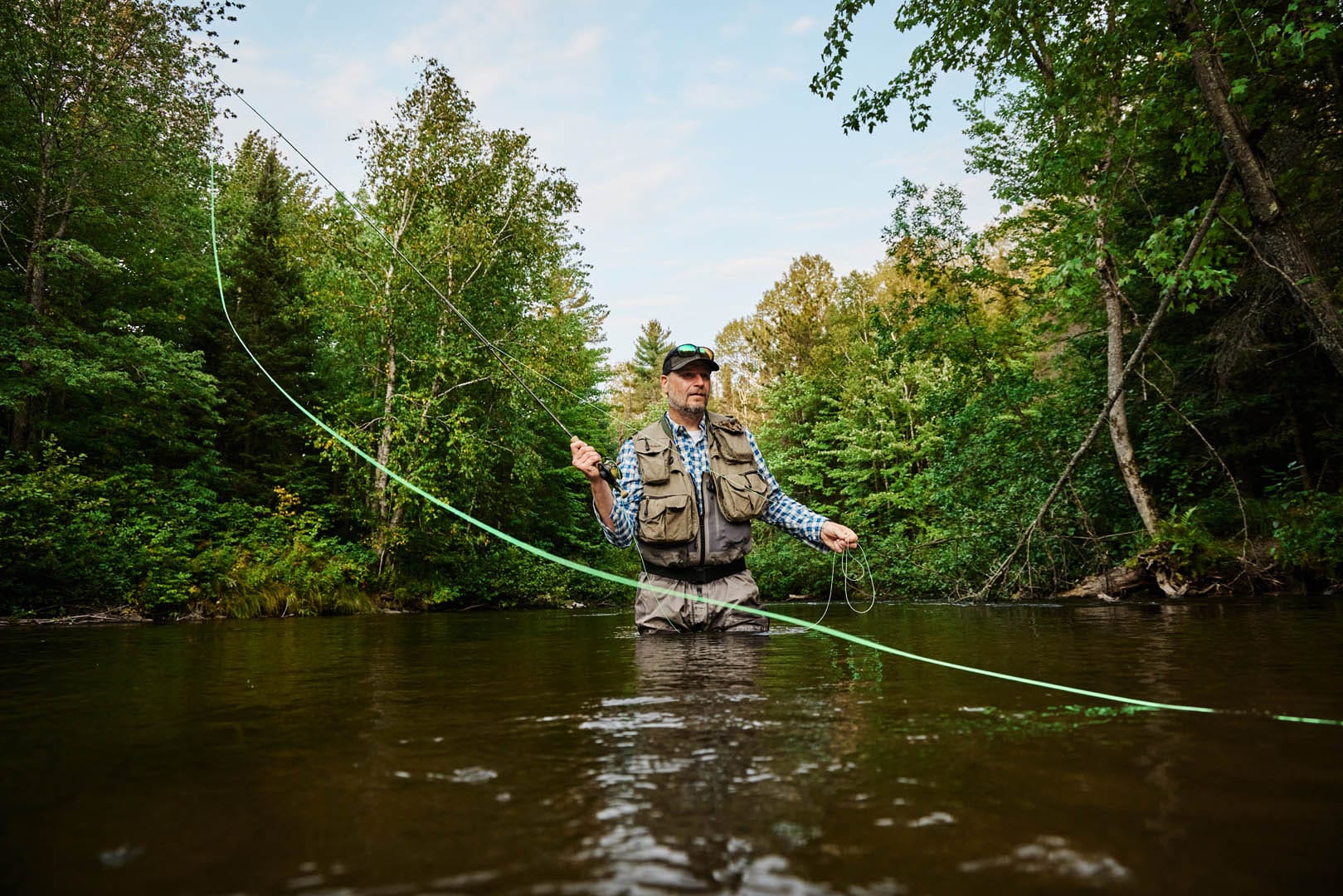 A man fly-fishing in a deep, dark river. He is wearing waders that allow him to be in the water without getting wet. The water is at his groin level. The trees along the river are bright green.