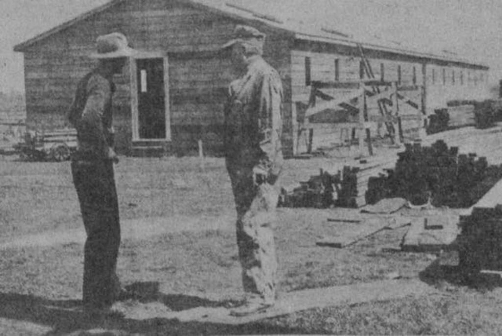 A very old, black and white photo, of two men talking in a lumber yard near a building.