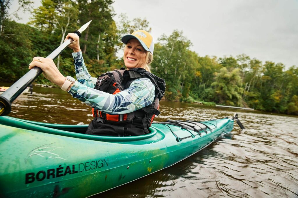 A close up photo of a smiling woman with blonde har and a yellow had is kayaking in a bright green kayak.