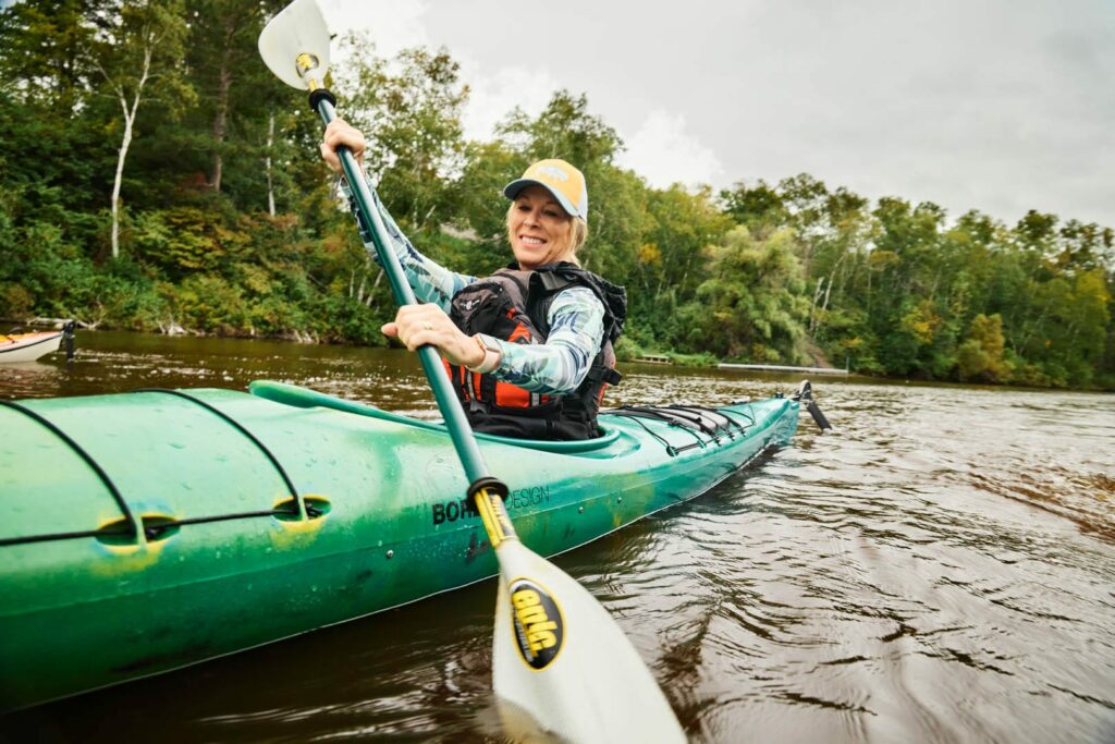 A close up photo of a smiling woman with blonde har and a yellow had is kayaking in a bright green kayak.