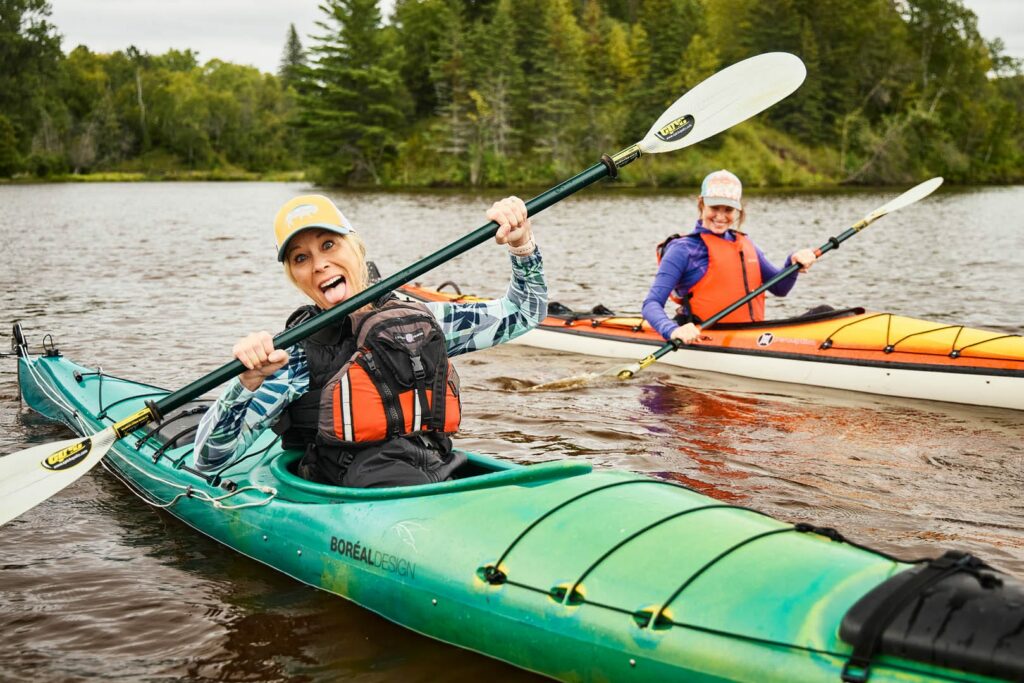 Two women are kayaking as they look at the camera happily.