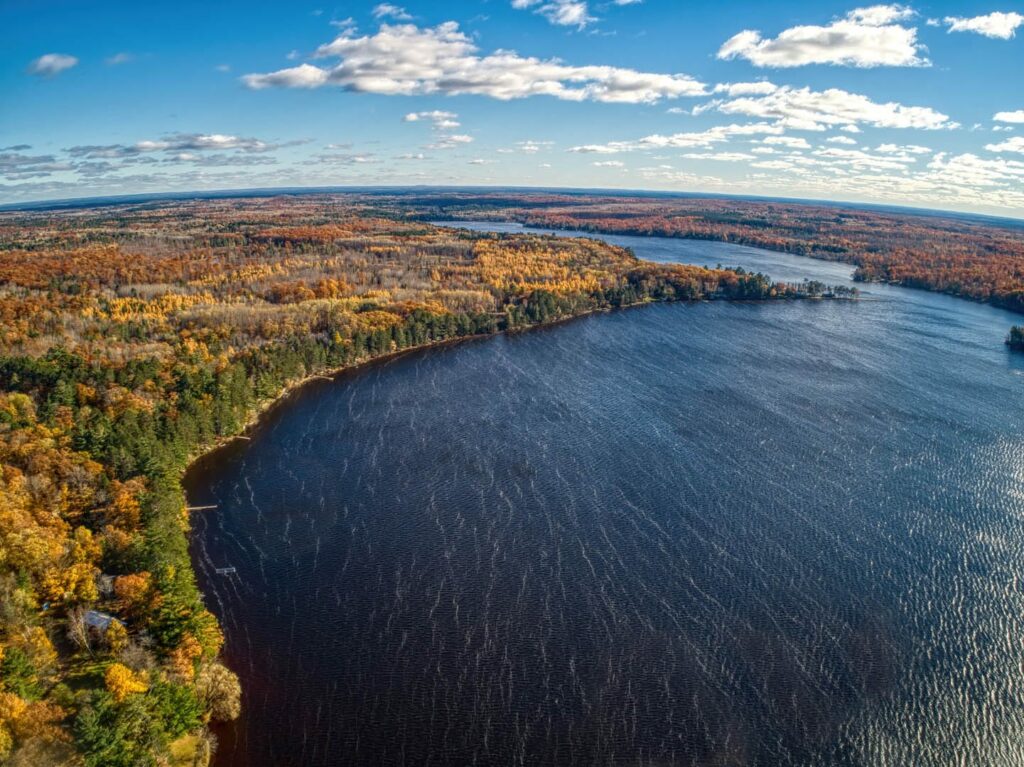 A birds-eye view of a lake with dark blue water and autumn colored forest around its shoreline.