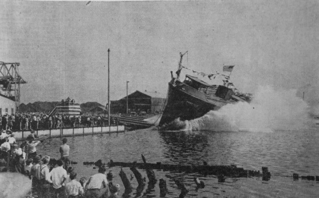 A black and white old photo of a ship being slid into the water with a large splash.
