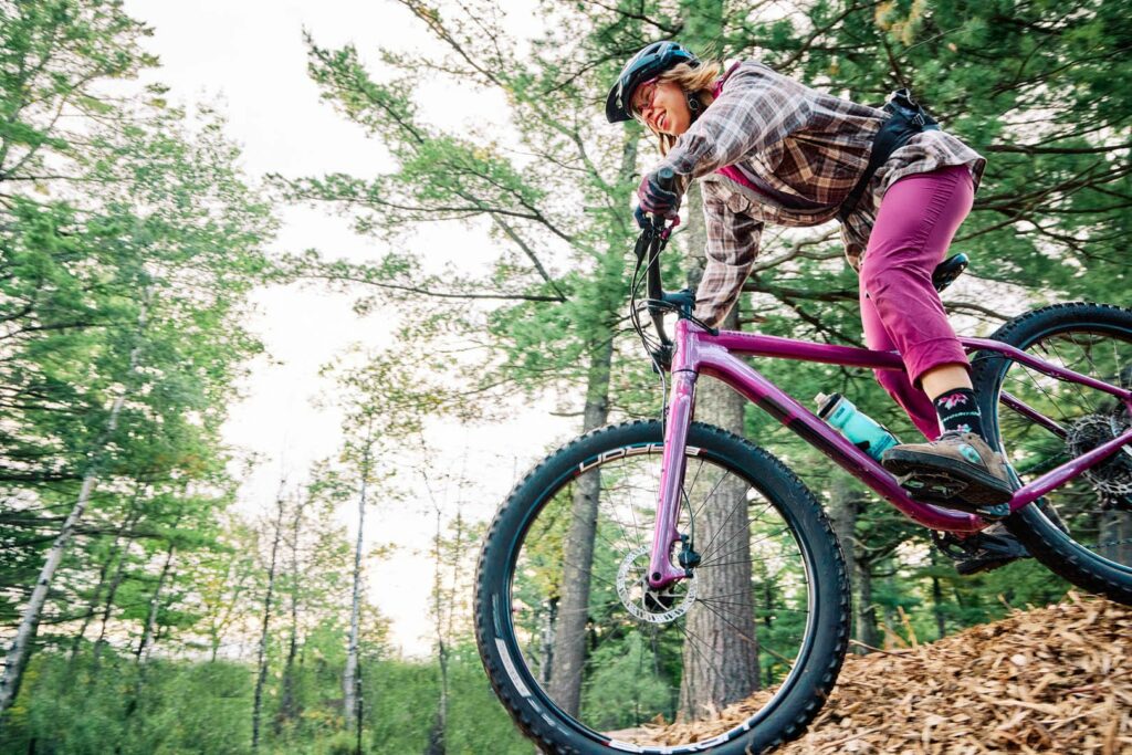 A female mountain biker rides her purple bike through the woods.