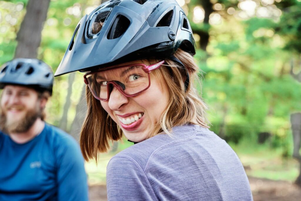A young woman with dark pink colored rims on her glasses and with matching lipstick turns to the camera and makes a funny face. She is wearing a blue helmet.