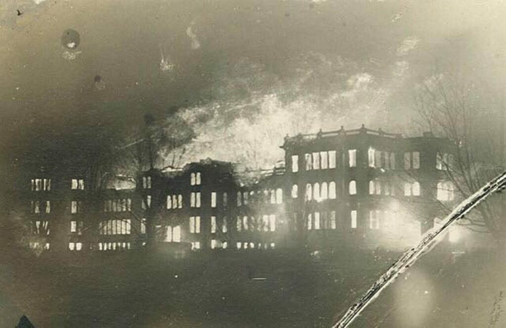 An very old sepia-toned image of a large school burning with flames coming out of the top and every window.