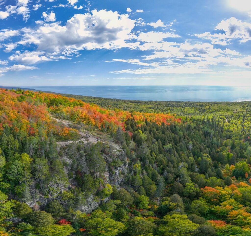 A hillside view of Lake Superior with bold autumn colored trees from dark green to blaze orange. Lake Superior is very blue and way off in the distance.