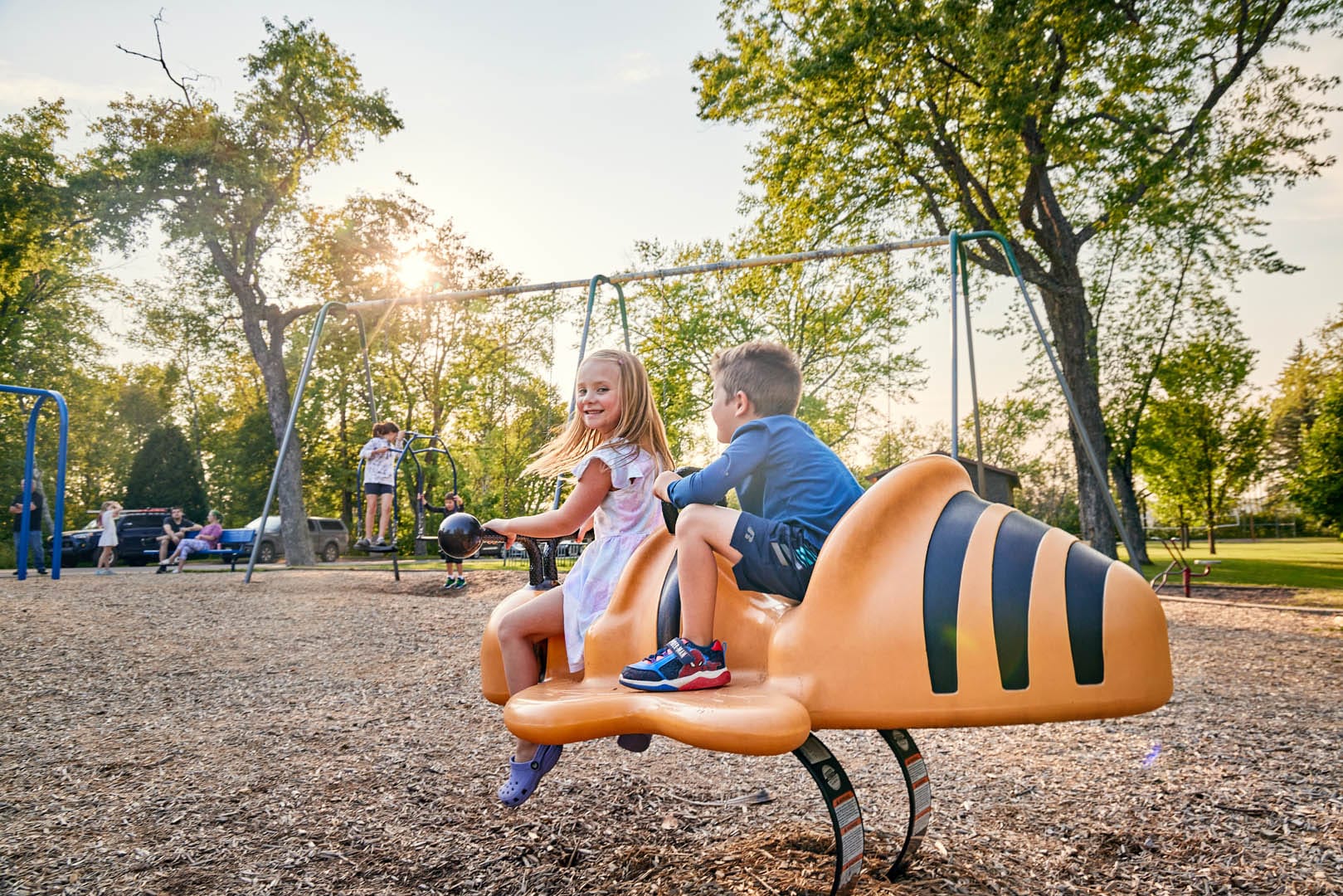 A young boy and a young girl playing on a large bumblebee at a park. There is a swing set behind them.
