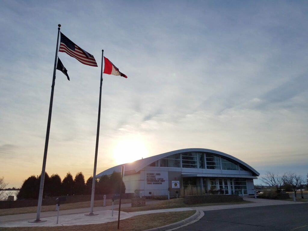 Exterior photo of the Richard I. Bong Veterans Historical Center with the sun shining behind the building. There are three flags blowing in the wind in front of the building.