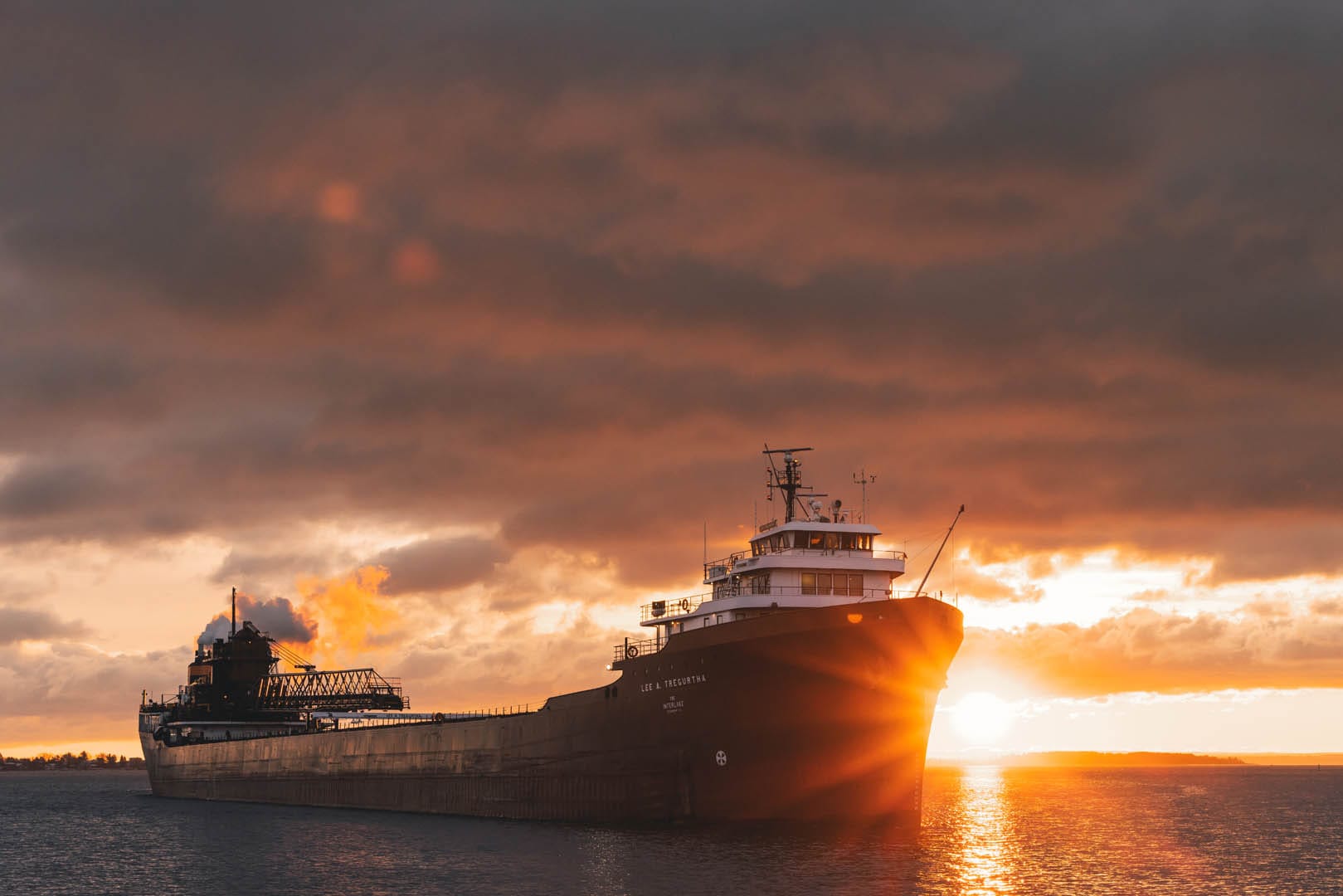 A laker with a sunrise in the background. The entire photo has an orange effect from the color of the sunrise. The ship is traveling as there is smoke coming out of the chimney in the back.