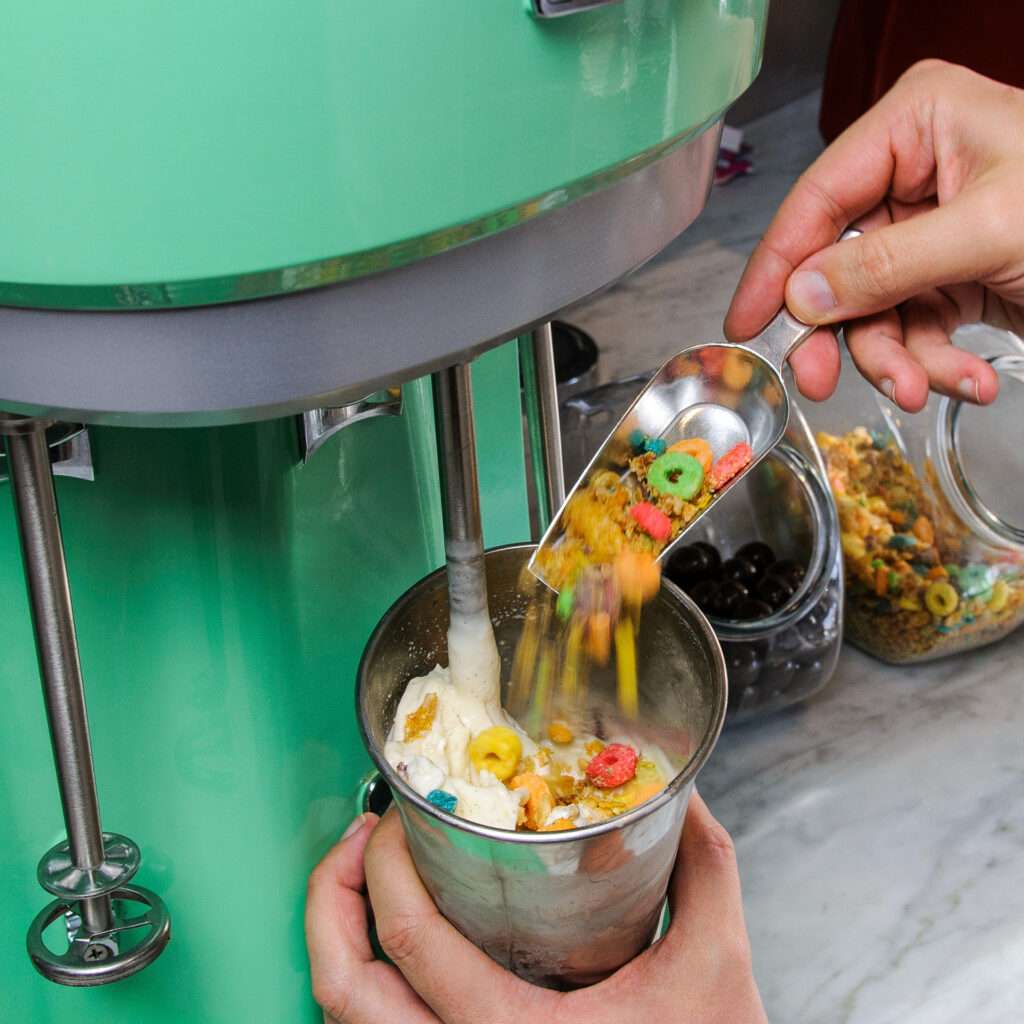 A green old fashioned malt maker. A person is holding the metal cup under the mixer and adding candy and cheerios to the ice cream mixture.