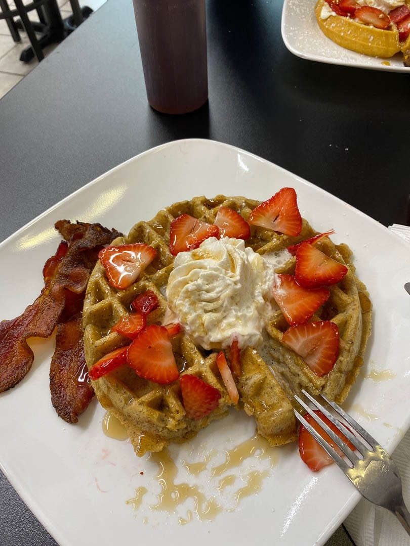 A large Belgian waffle topped with whipped cream, strawberries, and a side of bacon.