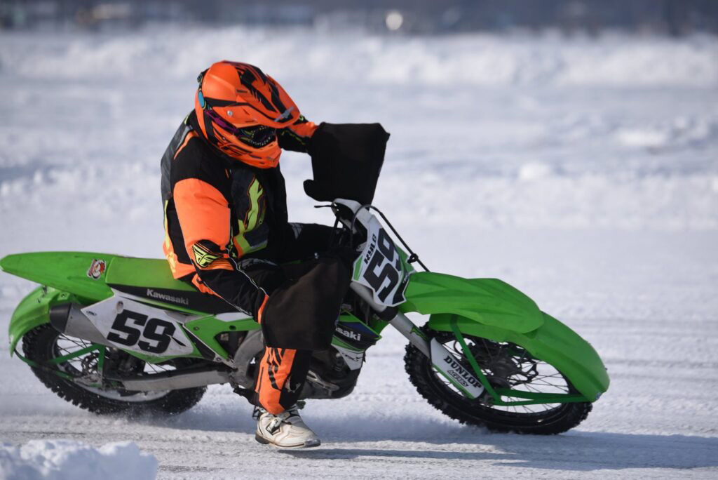 A green motorized dirt bike and rider in black and orange clothing and orange helmet on an icy and snowy surface.