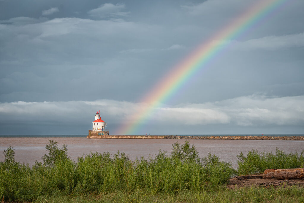 A rainbow arcs directly into the Superior Lighthouse with dark stormy clouds in the distance and bright green vegetation in the foreground. The lighthouse is white with a red roof.