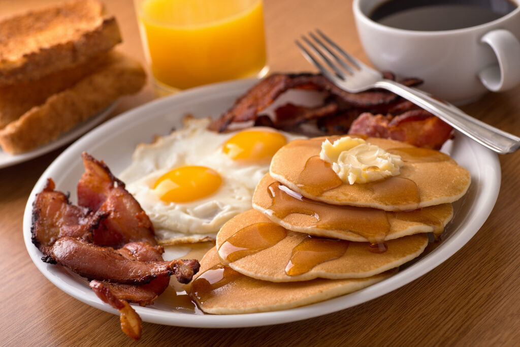 A plate of breakfast with four pancakes covered in syrup, strips of bacon, and two eggs sunny side up.