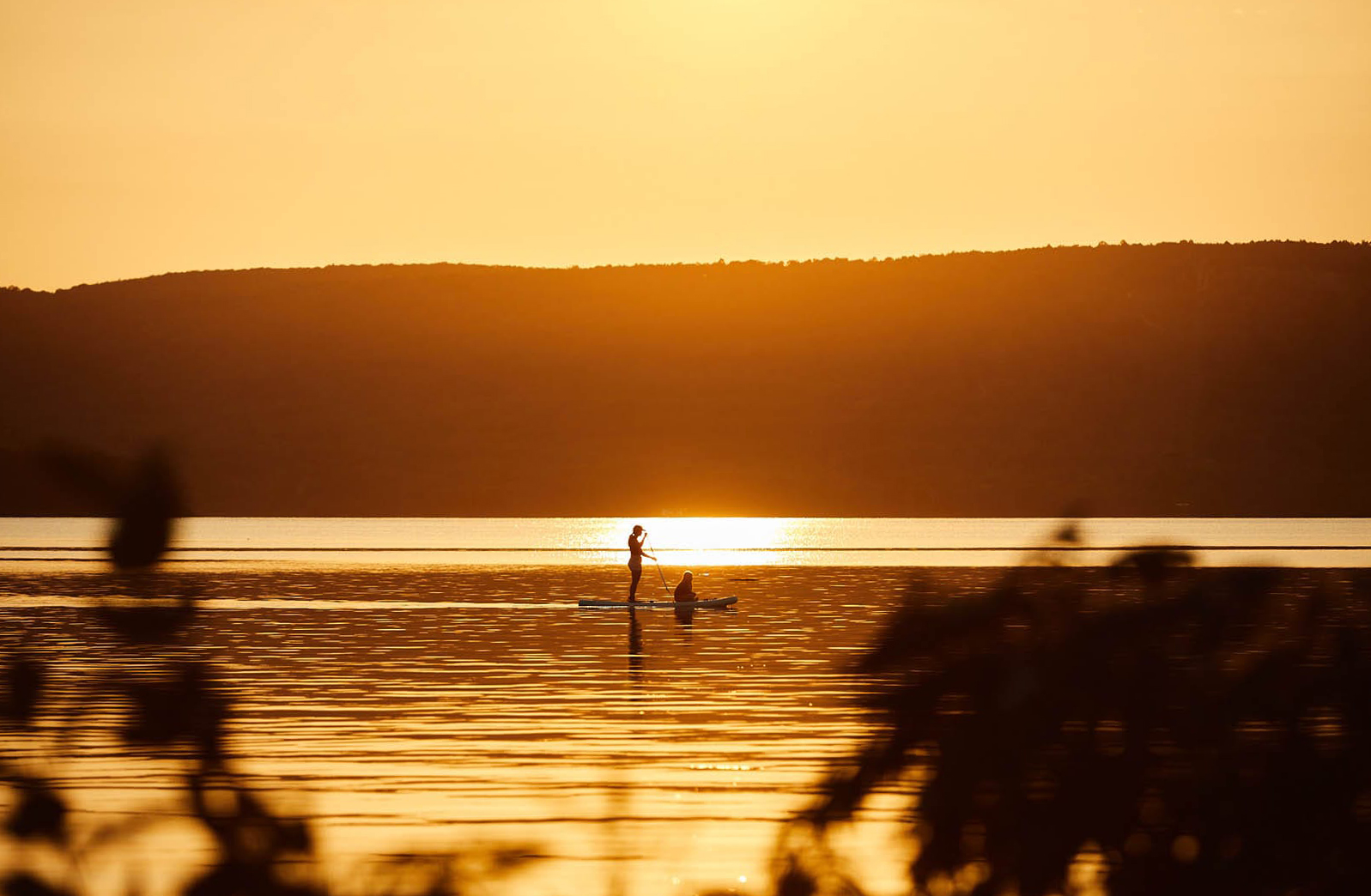 A woman standing on a paddle board with someone sitting on the front. The light is very warm and golden.