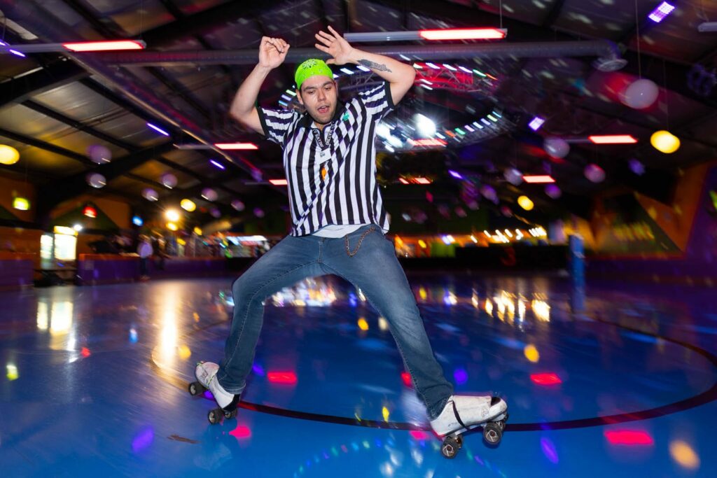 A roller skater in a black and white striped shirt, doing a trick on his skates on the roller rink.