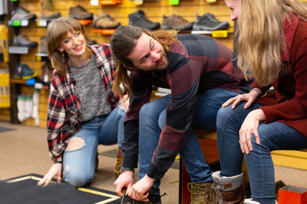 Three people at a shoe store, chatting to each other and trying on hiking boots.