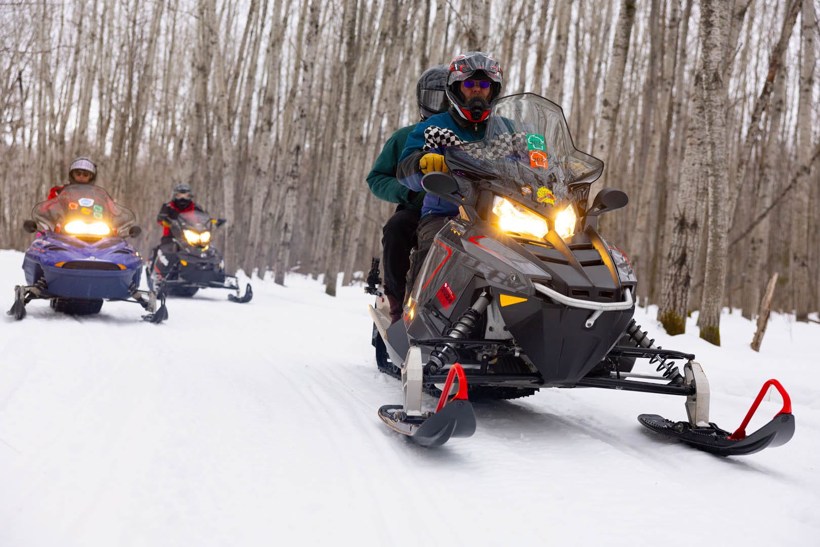 Three snowmobilers with passengers traveling through the woods in later part of the day with their headlights on.