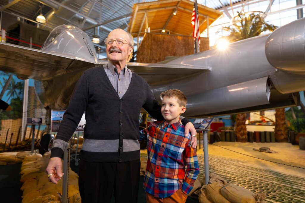 An older man with his arm around his grandson, looking at World War Two memorabilia.