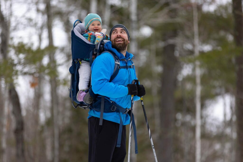 A father and his baby are cross country skiing. He has the baby girl on his back in a backpack. They've stopped to have a little chuckling moment as he looks back at her and she looks at the camera.
