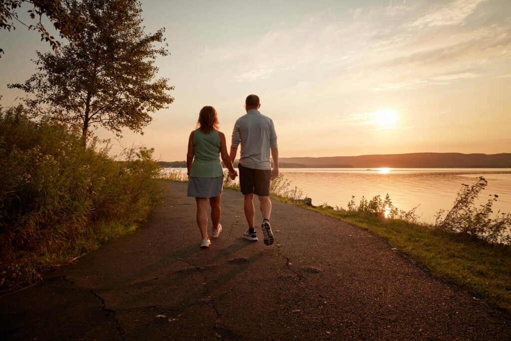A couple walking and holding hands on a trail along a body of water. The sun is setting behind them.
