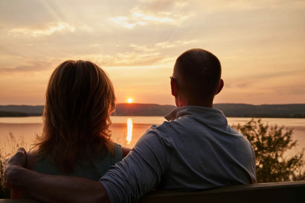 From behind, two people sitting on a bench overlooking a very warm sunset along a large body of water also known as the St. Louis River. He has his arm around her.