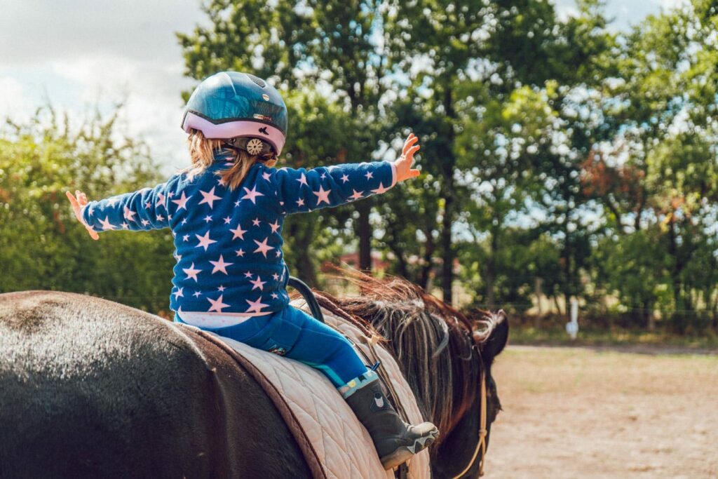 A toddler in a starry-felted jacket and helmet rides a small horse. Her arms are extended out like 