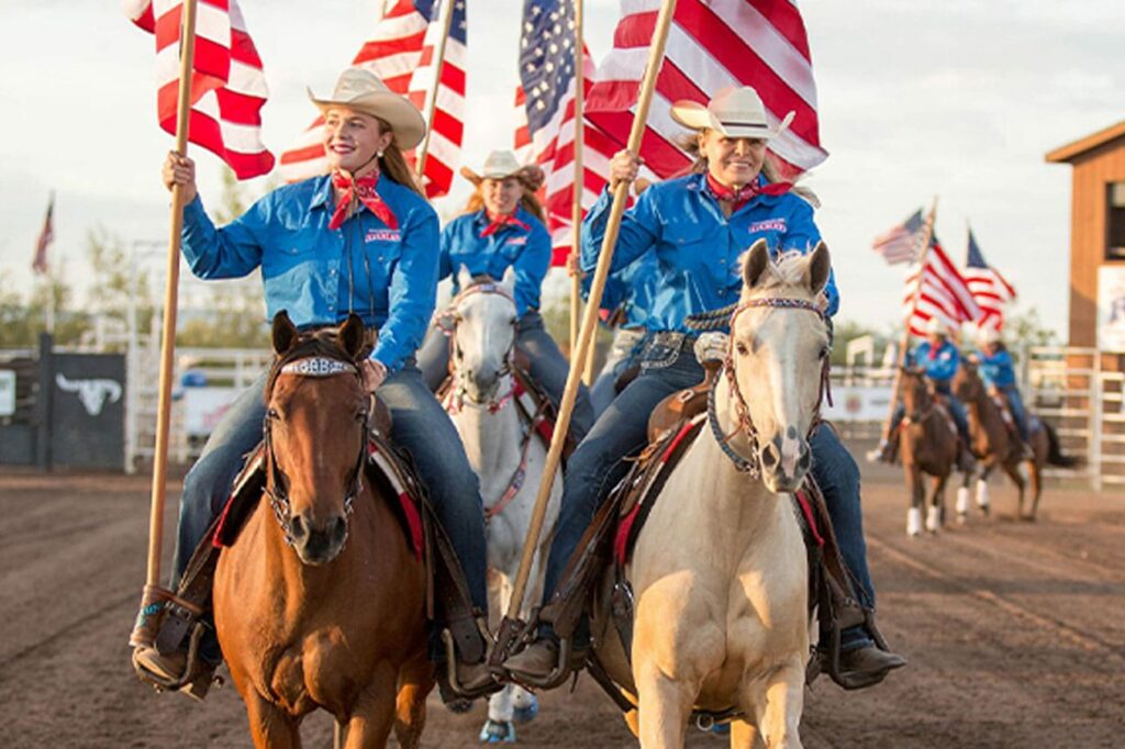 Three women in blue shirts and jeans riding horses each holding the American flag at a rodeo.