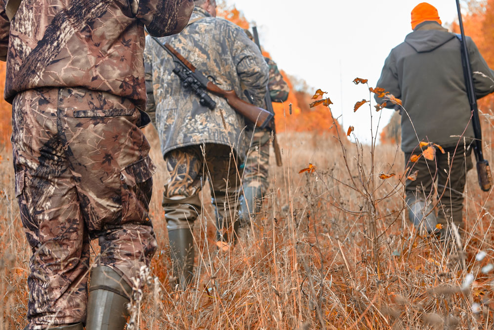Four guys wear cameo and hold guns with scopes as they head out into a field to hunt.