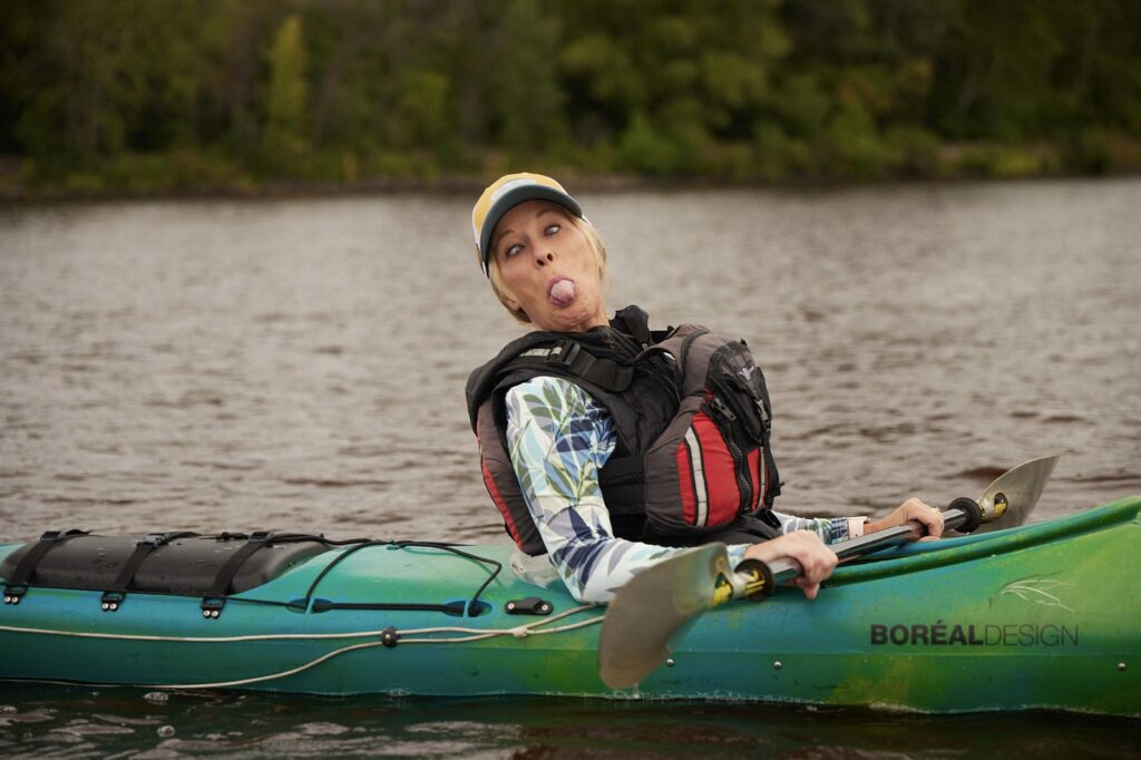 A blonde woman in a kayak is making a funny face at the camera. Her tongue is sticking out and her eyes are crossed.