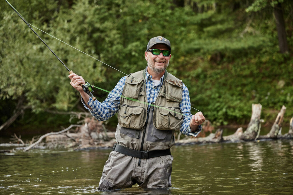A smiling man is up to his thighs in a river. He is holding a fishing pole.