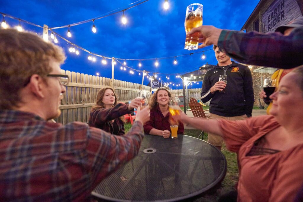 A group of people smile and raise their beers for a cheers. They are on the Earth Rider Patio at night.