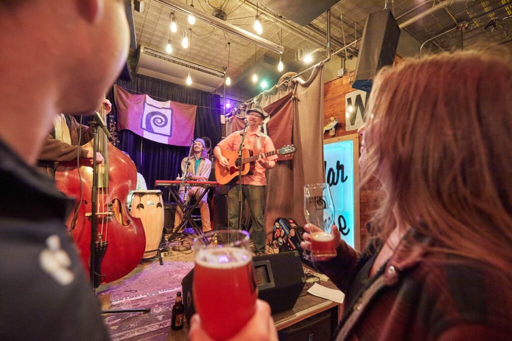Group of people drinking beer at the cedar lounge while a band with a guitar, double bass, and keyboard perform on stage.