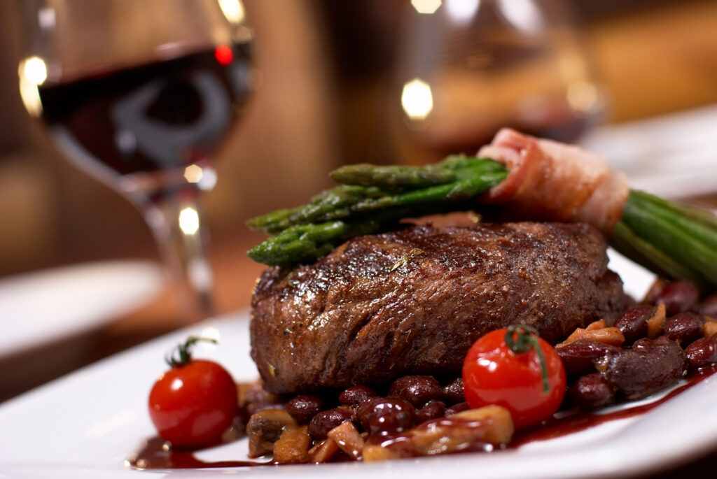 A thick cooked steak with bacon wrapped asparagus on top, two cherry tomatoes on the plate and a glass of red wine in the background.