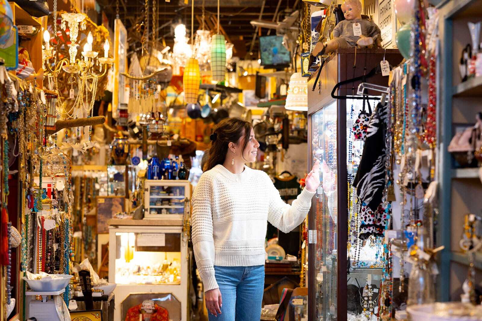 A woman is looking through a crowded and eclectic antique store. There are even antiques hanging form the ceiling.