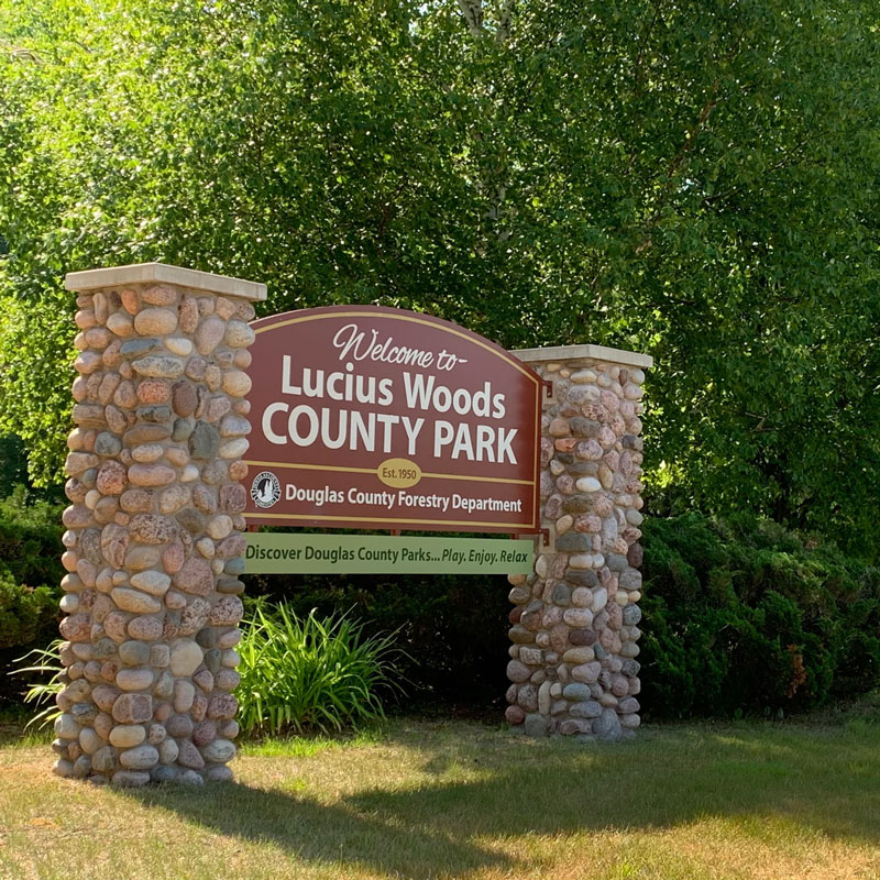 An outdoor sign surrounded by trees. The sign is for Lucius Woods County Park.