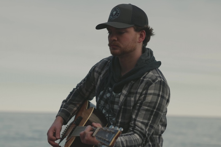 A younger man playing the guitar next to the water.