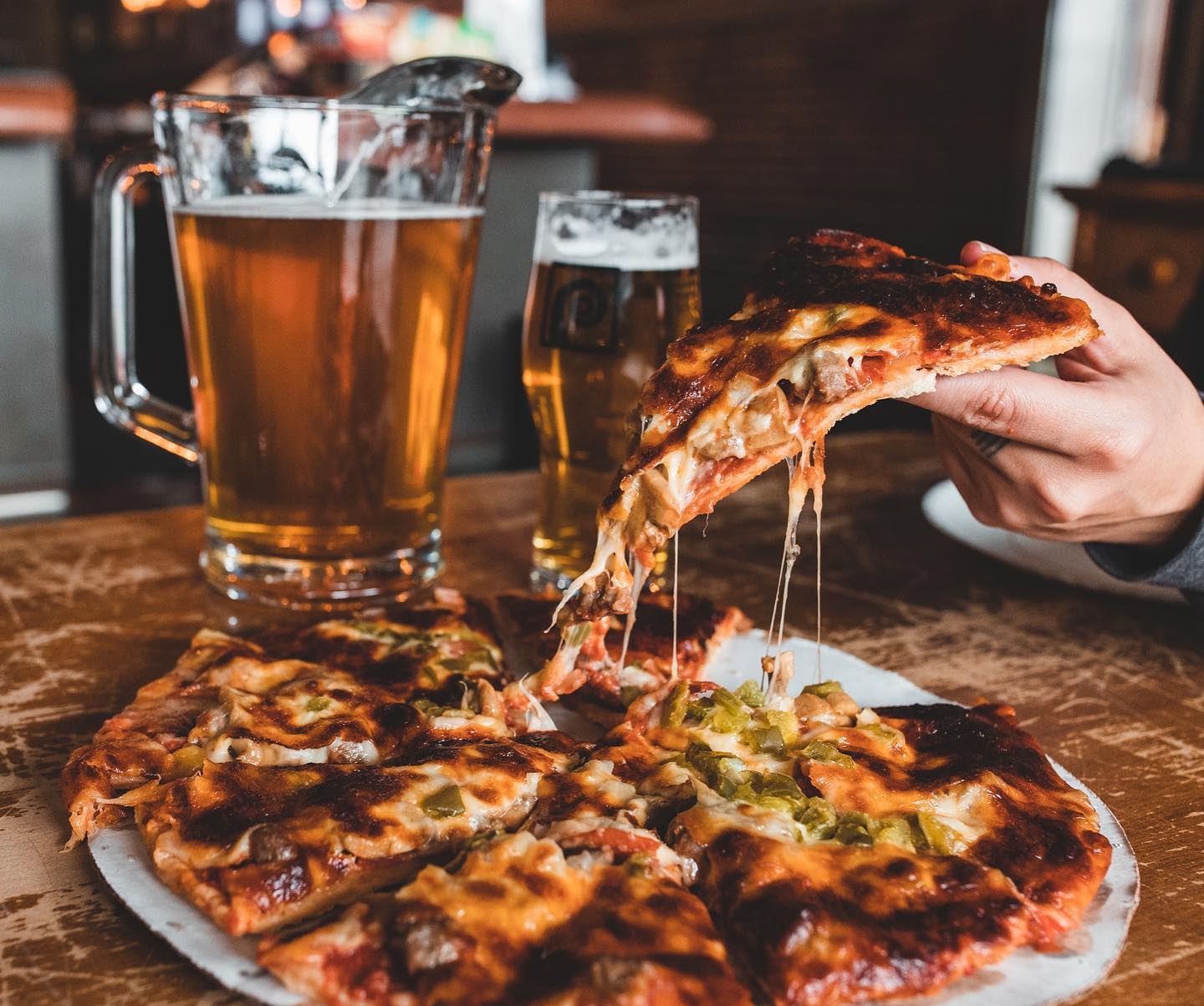 A pizza loaded with toppings on a table with a pitcher and a pint of beer.