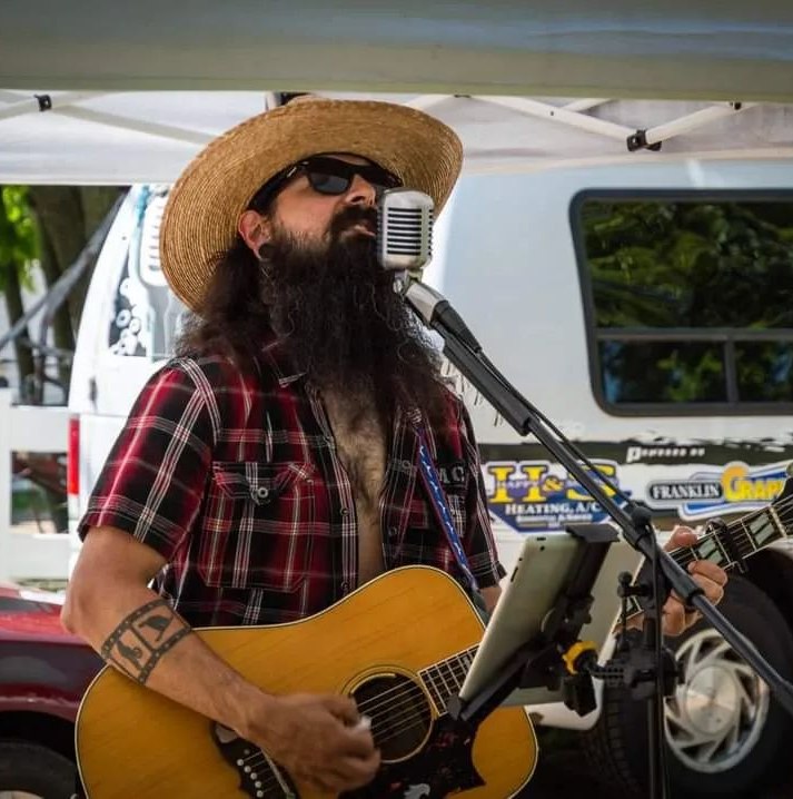 A middle aged man in a hat and sunglasses with a large beard plays guitar and sings into a microphone